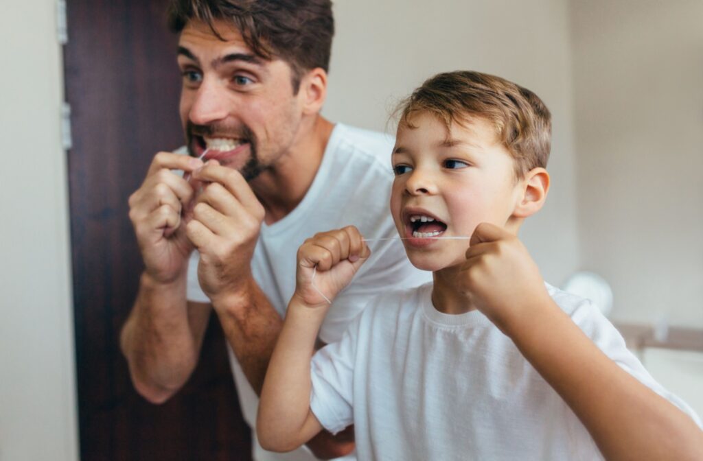 Smiling father and young son flossing their teeth together in front of a bathroom mirror.