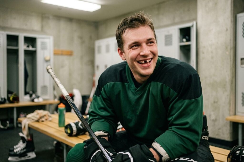 Smiling adult hockey player missing a front tooth sits on a locker room bench in a green jersey, holding a hockey stick.